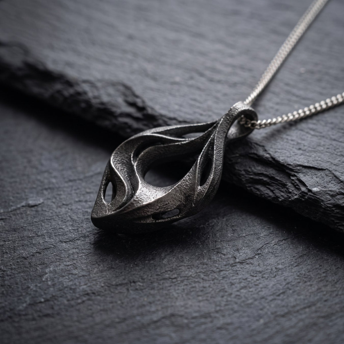 Illustrative close view of the recognizable UNDA pendant resting on slate, showing its textured titanium surface